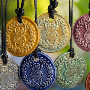 Può includere: Una collezione di ciondoli circolari in ceramica appesi a cordini neri. Ogni ciondolo presenta uno scarabeo modellato in rilievo al centro di una corona circolare di foglie e piccoli fiori. I ciondoli sono smaltati in vari colori tra cui blu, giallo, verde e viola.