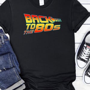 Może przedstawiać: Czarny T-shirt z kolorową grafiką z napisem "Back to the 80s" w retro czcionce.