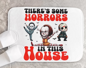 Puede incluir: Alfombra de baño blanca con un gráfico rojo y blanco de tres personajes de películas de terror en una pose de baile. El texto dice "There's some horrors in this house".