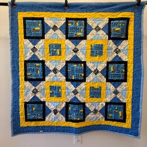May include: A blue, yellow, and black quilt with a repeating pattern of squares. The squares feature images of yellow construction vehicles and the text "TONKA TOUGH". The quilt is hanging from a black rod with clothespins.