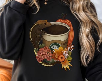 Könnte beinhalten: Schwarzes Sweatshirt mit einem Design einer Cappuccino-Tasse, einem Gecko, einem Kürbis und Herbstlaub. Auf der Tasse steht das Wort "cappy". Das Design hat orange, braune und gelbe Farben, ideal für die Herbstsaison.