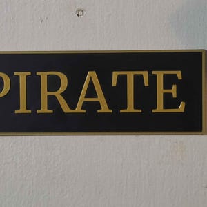 May include: A rectangular sign with the word "PIRATE" in gold lettering on a black background. The sign has a gold border and is mounted on a white wall.