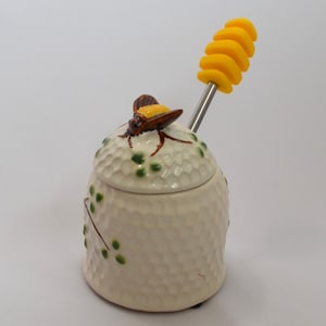 May include: Cream-colored ceramic honey pot with a textured surface and a bee-shaped lid. A yellow honey dipper is inserted into the pot. Green leaf and berry accents decorate the side of the pot.
