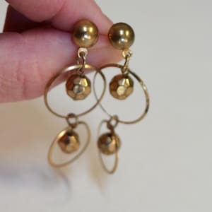May include: A pair of gold-toned dangle earrings featuring a stud top with a smooth sphere, followed by a larger circular hoop containing a faceted spherical bead, and a smaller circular hoop at the bottom with another faceted spherical bead.