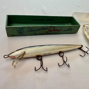 May include: A silver and white Rapala fishing lure with three treble hooks and a clear plastic lip, labeled Finland. The lure is accompanied by its original green cardboard box with the text Floating Model and a printed instruction paper.
