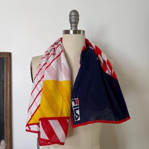 May include: A rectangular scarf featuring a color-blocked design with sections of white and red stripes, a solid yellow diagonal panel, and a large navy blue section. The scarf includes the white FILA logo with red and blue lettering on the navy side.