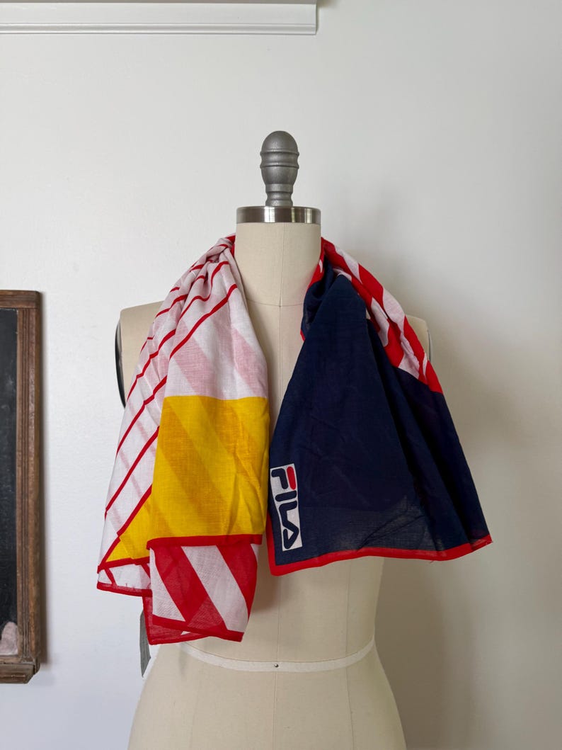 May include: A rectangular scarf featuring a color-blocked design with sections of white and red stripes, a solid yellow diagonal panel, and a large navy blue section. The scarf includes the white FILA logo with red and blue lettering on the navy side.