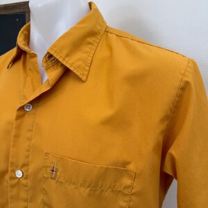 May include: Mustard yellow button-down shirt with a chest pocket and a small embroidered design. The shirt has a classic collar and long sleeves. The fabric appears to be a lightweight cotton or blend.