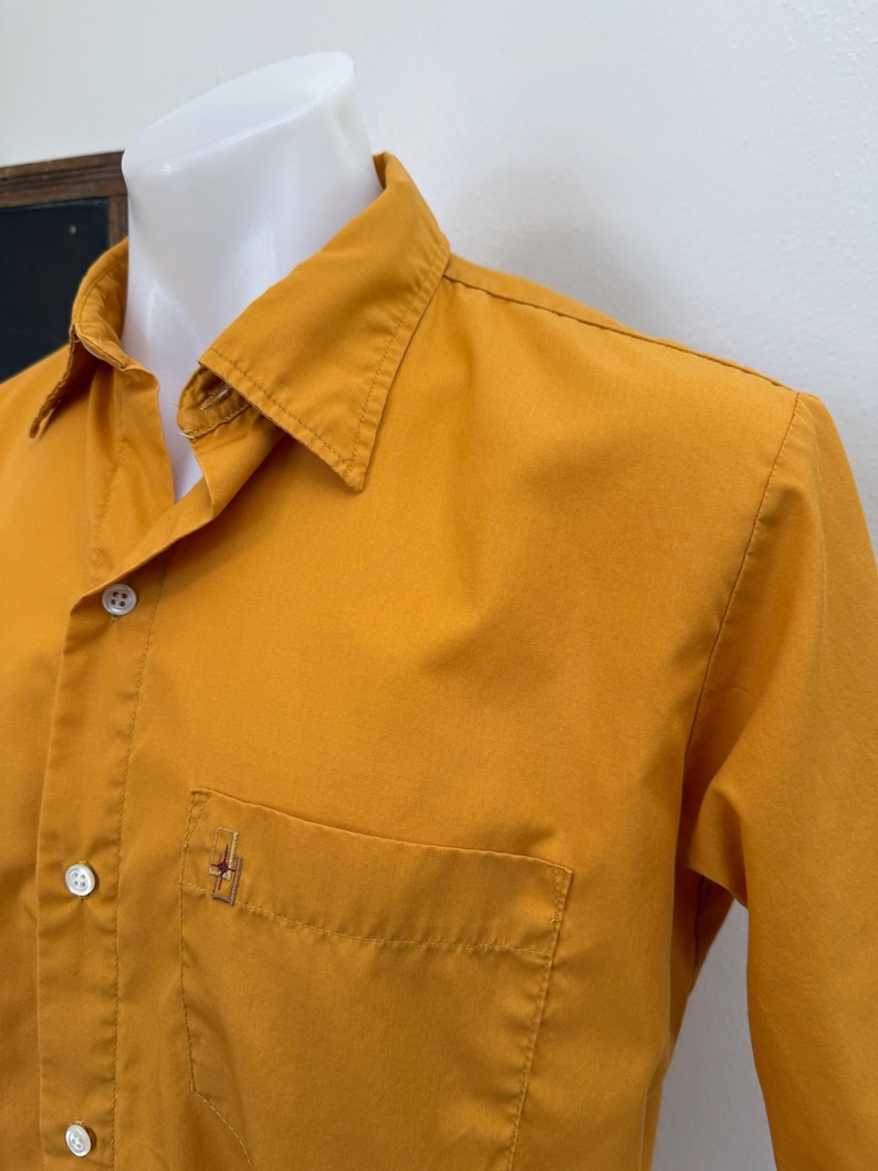 May include: Mustard yellow button-down shirt with a chest pocket and a small embroidered design. The shirt has a classic collar and long sleeves. The fabric appears to be a lightweight cotton or blend.