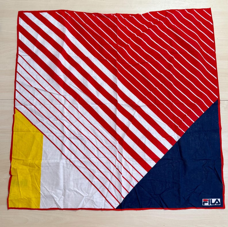 May include: A square bandana featuring a diagonal pattern of red and white stripes across the top and center. The bottom left corner has a solid yellow block, while the bottom right corner is a solid navy blue triangle with the FILA logo in white.