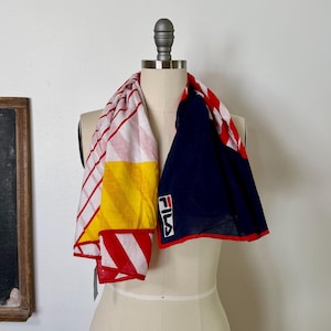 May include: A Fila scarf featuring a color-blocked design with a white section displaying red diagonal stripes, a solid yellow panel, and a large navy blue section. The Fila logo is printed in white, red, and navy on the navy blue fabric.