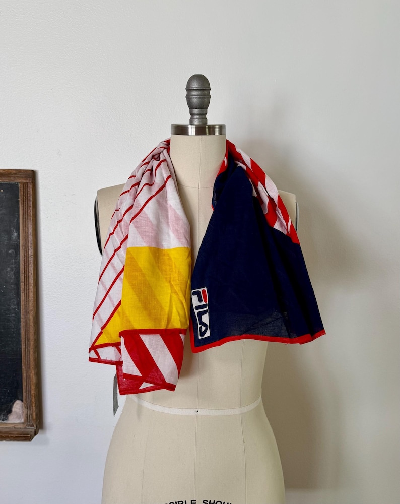 May include: A Fila scarf featuring a color-blocked design with a white section displaying red diagonal stripes, a solid yellow panel, and a large navy blue section. The Fila logo is printed in white, red, and navy on the navy blue fabric.