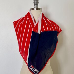 May include: A square scarf featuring a bold red and white vertical striped pattern on one side and a solid navy blue section on the other. The scarf displays the white FILA logo with a red and blue accent bar printed on the navy blue corner section.