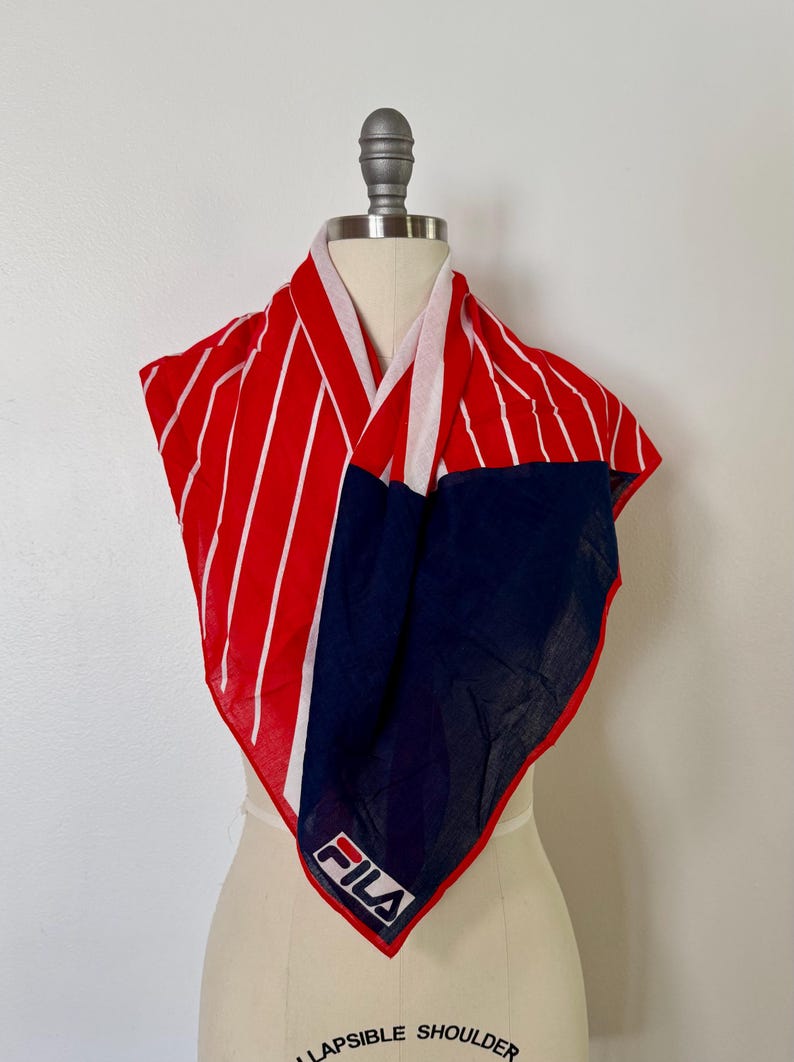 May include: A square scarf featuring a bold red and white vertical striped pattern on one side and a solid navy blue section on the other. The scarf displays the white FILA logo with a red and blue accent bar printed on the navy blue corner section.