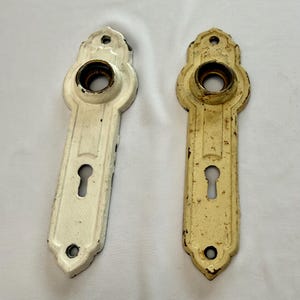 May include: Two elongated metal door plates with a central circular opening for a knob and a lower keyhole. One plate is finished in a clean white, while the other features a distressed, aged cream finish with visible signs of wear and oxidation.