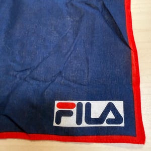May include: A dark blue fabric bandana featuring a bright red border along the edges. The bottom corner displays a rectangular white patch with the FILA brand logo printed in red and dark blue lettering, centered on the fabric surface.