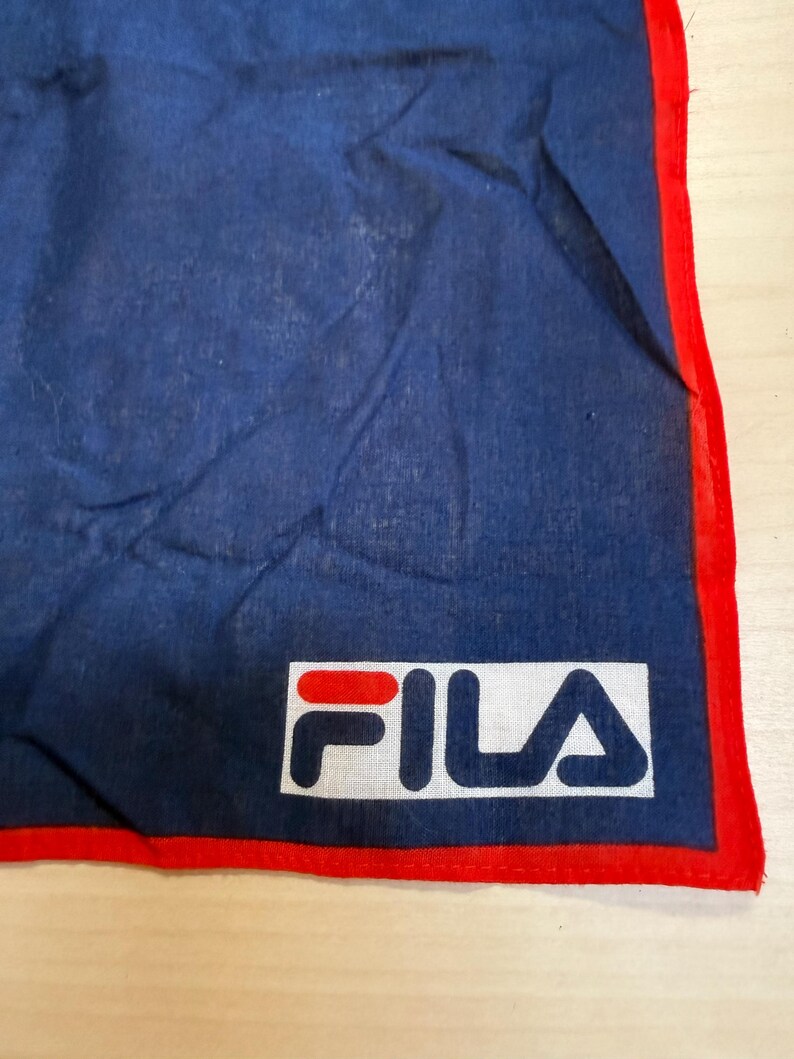 May include: A dark blue fabric bandana featuring a bright red border along the edges. The bottom corner displays a rectangular white patch with the FILA brand logo printed in red and dark blue lettering, centered on the fabric surface.