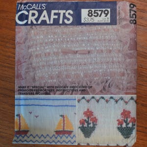 May include: A vintage McCall's Crafts pattern packet. The cover is blue with the text "McCall's Crafts" and the number 8579. The packet features examples of smocking and embroidered borders, including sailboats and floral designs.