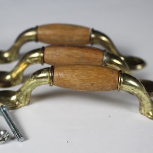 May include: Three cabinet pulls featuring a central barrel-shaped wooden grip with a light brown finish, flanked by polished brass metal ends and mounting plates. The set includes matching circular brass base plates and three silver-coloured metal screws.