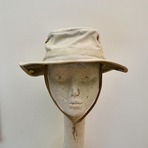May include: A beige bucket hat with a wide brim and a chin strap. The hat features small metallic buttons on the sides of the crown and the brim, allowing the sides to be folded up. The fabric appears to be a sturdy cotton or canvas material.