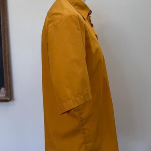 May include: Mustard yellow short-sleeved button-down shirt. The shirt is displayed on a white mannequin. The shirt has a collar and buttons down the front. The sleeves are short and end just above the elbow. The fabric appears to be cotton.