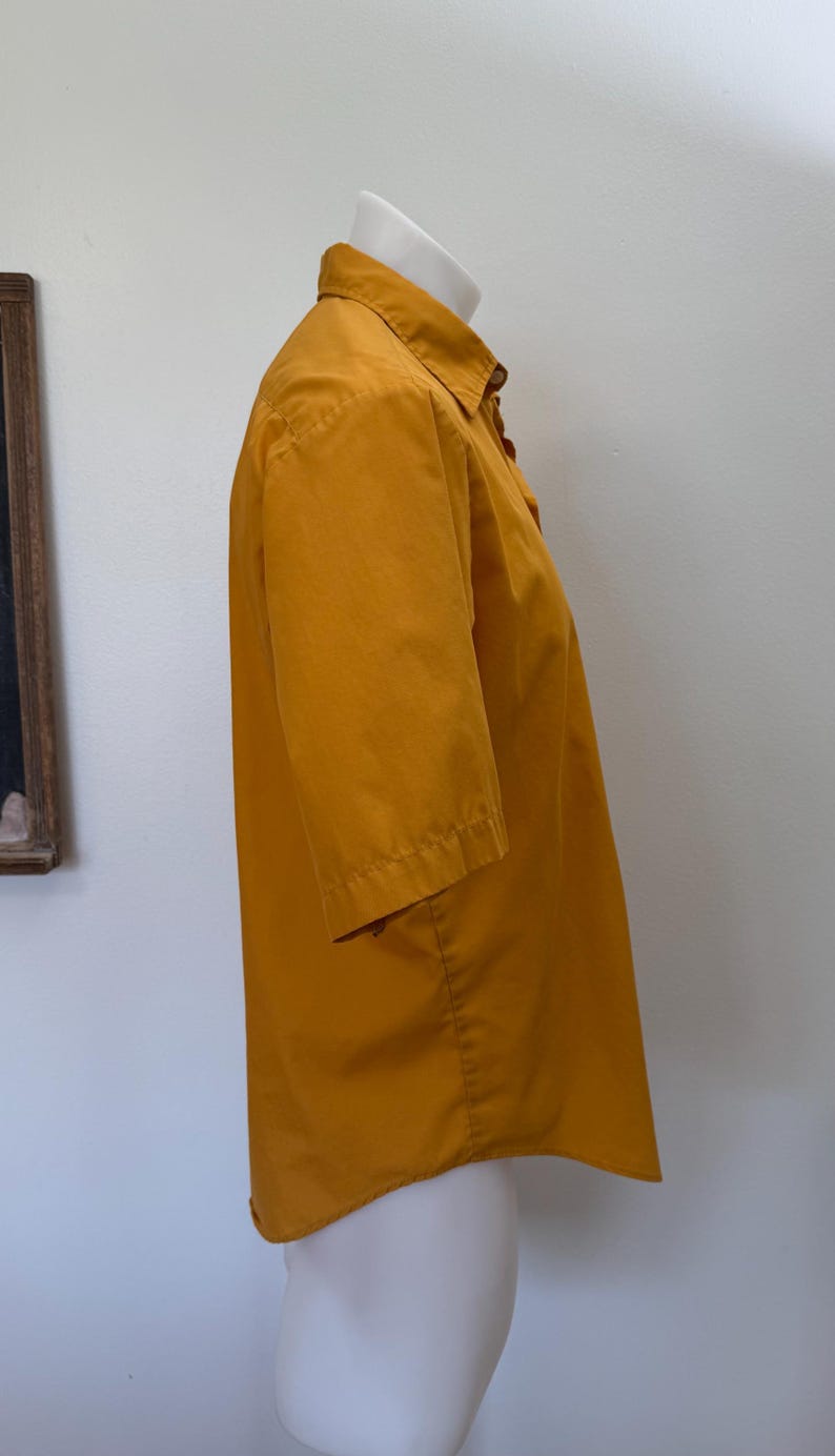 May include: Mustard yellow short-sleeved button-down shirt. The shirt is displayed on a white mannequin. The shirt has a collar and buttons down the front. The sleeves are short and end just above the elbow. The fabric appears to be cotton.
