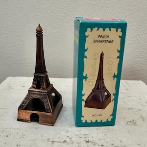 Puede incluir: Un sacapuntas de metal color cobre con forma de Torre Eiffel, acompañado de su caja de cartón original en color verde azulado y crema. La caja presenta el texto PENCIL SHARPENER, NO.165 y un texto escrito a mano que dice Christmas 2001.
