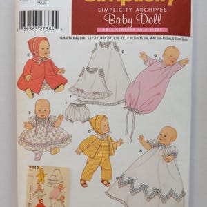 May include: A Simplicity sewing pattern envelope 5215 for baby doll clothes in sizes S, M, and L. The front features illustrations of dolls wearing various outfits including a red coat, a floral dress, a yellow gingham set, and a long white gown.