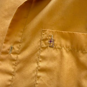 May include: Close-up of a mustard-yellow button-down shirt. The shirt has a small pocket on the chest with a decorative embroidered design. The fabric has a subtle sheen, and the stitching is visible.