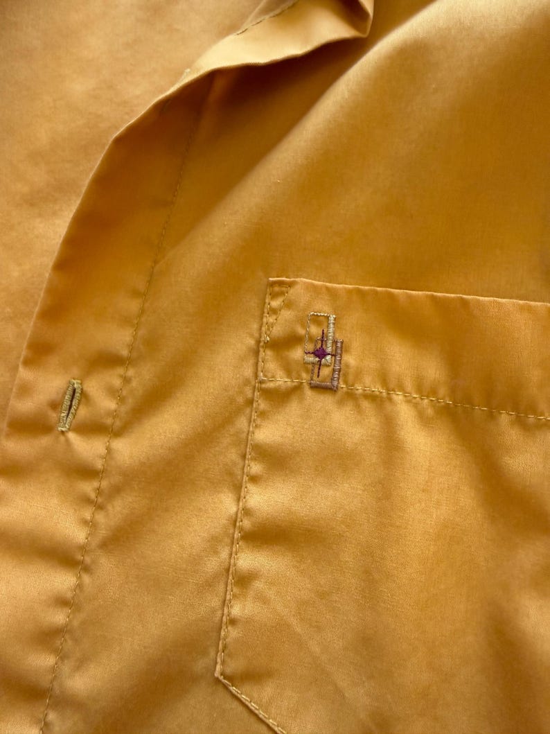 May include: Close-up of a mustard-yellow button-down shirt. The shirt has a small pocket on the chest with a decorative embroidered design. The fabric has a subtle sheen, and the stitching is visible.