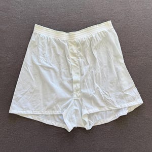 May include: White boxer shorts with an elastic waistband and button closure. The shorts are made of a lightweight, slightly wrinkled fabric. The garment is laid flat on a gray surface.