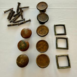 Pode incluir: Uma variedade de componentes de hardware antigos, incluindo botões de latão, puxadores quadrados e parafusos. Os botões são redondos com um topo ligeiramente abobadado. Os puxadores quadrados têm um design geométrico simples. Os parafusos são de cor bronze.