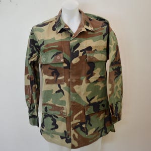 Könnte beinhalten: Hemd mit Tarnmuster, mit langen Ärmeln und Knöpfen. Das Hemd hat ein klassisches Design im Militärstil mit mehreren Taschen und einem Kragen. Die Farbpalette umfasst Grün-, Braun- und Schwarztöne, wodurch ein Waldtarnungseffekt entsteht.