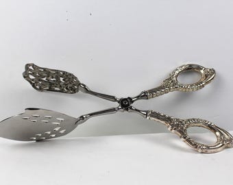 May include: A pair of silver-toned metal serving tongs featuring ornate, embossed handles with circular finger loops. One end has a solid, pointed, perforated blade, while the other end features a decorative, openwork filigree design.
