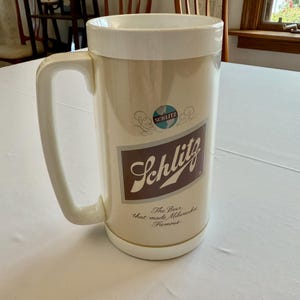Op de afbeelding: Crèmekleurige bierpul met een witte handgreep en rand. De pul heeft het Schlitz-logo in bruin en wit, met de tekst "The Beer that made Milwaukee Famous."