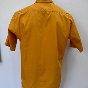 May include: Mustard yellow short-sleeved shirt with a classic collar. The shirt is displayed on a white mannequin. The back of the shirt is visible, showing the yoke and the curved hemline. The fabric appears to be a smooth, woven material.