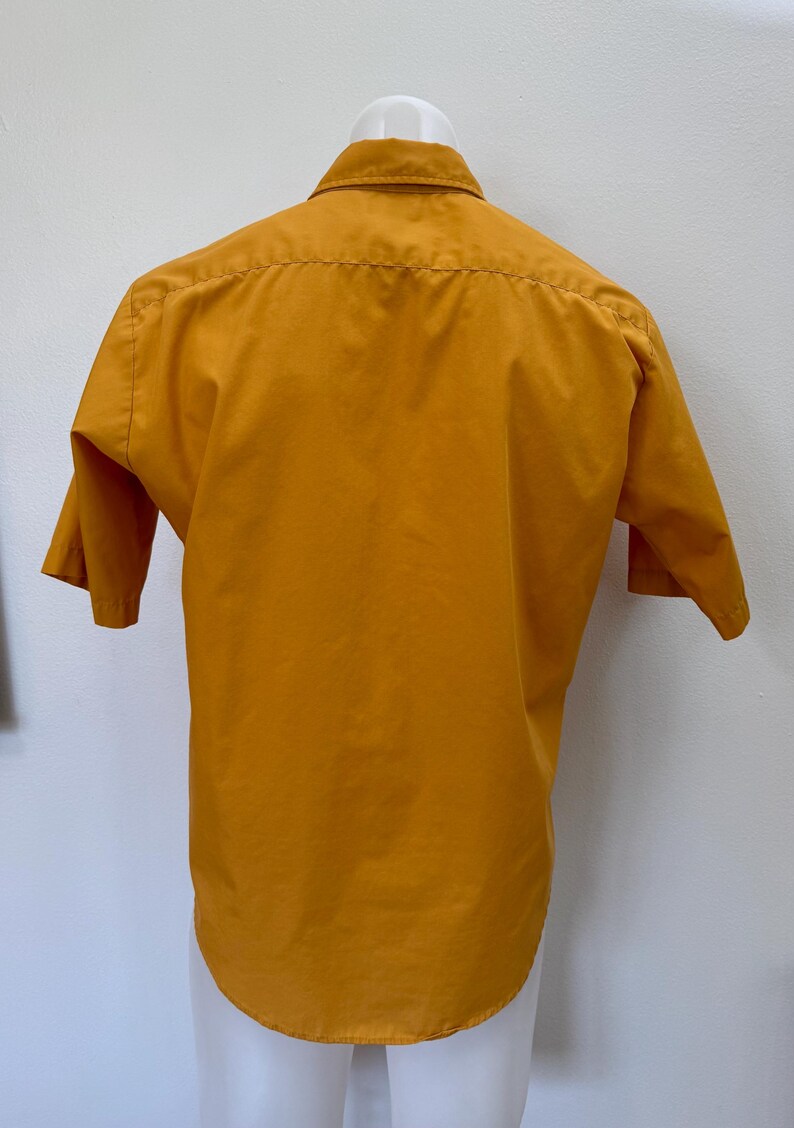 May include: Mustard yellow short-sleeved shirt with a classic collar. The shirt is displayed on a white mannequin. The back of the shirt is visible, showing the yoke and the curved hemline. The fabric appears to be a smooth, woven material.