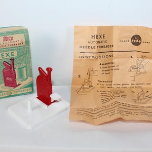 May include: A Hexe automatic needle threader with a red metal mechanism on a white plastic base, accompanied by its original teal and white cardboard box and a tan paper instruction sheet featuring diagrams and the text Hexe Automatic Needle Threader.