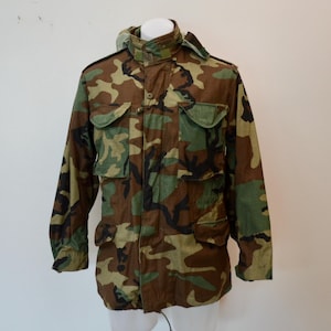 Könnte beinhalten: Tarnmusterjacke mit Kapuze und mehreren Taschen. Die Jacke hat ein Muster aus Grün, Braun und Schwarz. Sie wird auf einer Schaufensterpuppe vor einem schlichten weißen Hintergrund gezeigt. Die Jacke hat eine Knopfleiste vorne und einen Kordelzug am Saum.