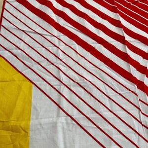 May include: A textile fabric featuring a pattern of bold red diagonal stripes on a white background, with a solid block of bright yellow color visible along the left edge. The material displays a visible weave texture and soft folds throughout.