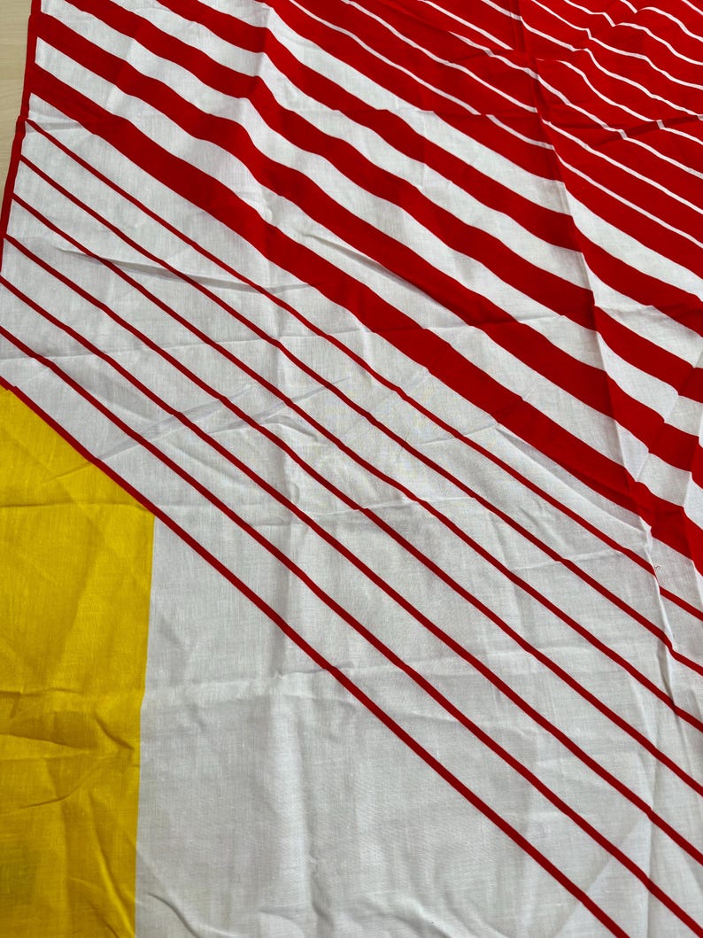 May include: A textile fabric featuring a pattern of bold red diagonal stripes on a white background, with a solid block of bright yellow color visible along the left edge. The material displays a visible weave texture and soft folds throughout.
