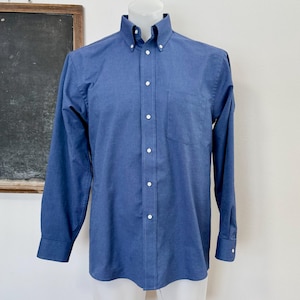 Op de afbeelding: Een blauw button-down overhemd met lange mouwen, een button-down kraag en een borstzak. Het overhemd wordt op een mannequin getoond. Het overhemd heeft witte knopen en manchetten. De achtergrond omvat een schoolbord en een witte muur.