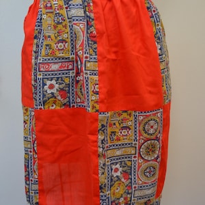 May include: A vintage apron with a patchwork design. The apron features a vibrant mix of patterns, including floral and geometric motifs in yellow, blue, and red. The apron has a solid red panel on the front and a blue patterned waistband.