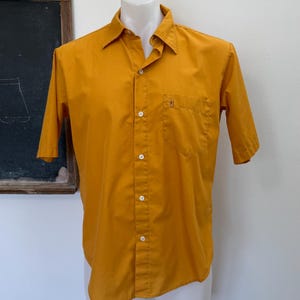 May include: Mustard yellow short-sleeve button-down shirt with a chest pocket. The shirt is displayed on a white mannequin. The shirt has a classic collar and white buttons. The background includes a chalkboard.
