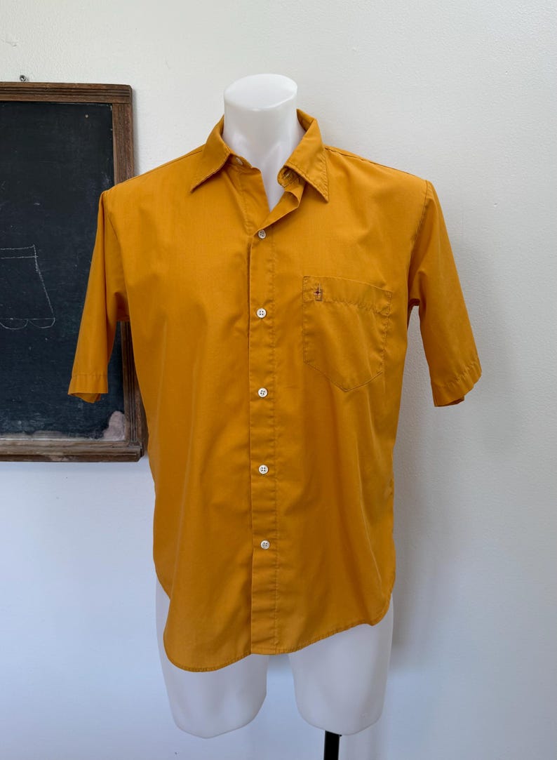 May include: Mustard yellow short-sleeve button-down shirt with a chest pocket. The shirt is displayed on a white mannequin. The shirt has a classic collar and white buttons. The background includes a chalkboard.