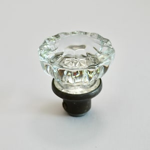 Op de afbeelding: Een heldere glazen kastknop met een facetachtig, bloemachtig ontwerp. De knop zit bovenop een donkere metalen basis. Deze decoratieve hardware is geschikt voor laden, kasten of meubels.