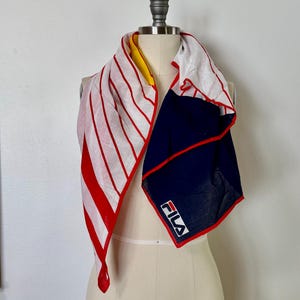 May include: A square scarf featuring a white background with red vertical stripes on one side and a solid navy blue section on the other. The scarf has a red border and displays the white FILA logo on the navy blue portion. A yellow accent is visible.