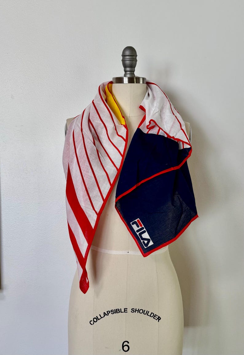 May include: A square scarf featuring a white background with red vertical stripes on one side and a solid navy blue section on the other. The scarf has a red border and displays the white FILA logo on the navy blue portion. A yellow accent is visible.