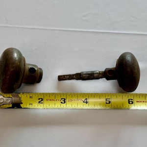 May include: Two antique brass door knobs with a dark, weathered patina. One knob is detached, while the other is attached to a square metal spindle. A yellow Stanley measuring tape is positioned below, showing the total length is about 7.5 inches.