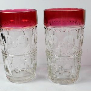 Puede incluir: Dos vasos de vidrio transparente con una banda de color rojo intenso en el borde superior. El cuerpo de cada vaso está decorado con un patrón en relieve de formas circulares y motivos florales grabados, creando una superficie texturizada.